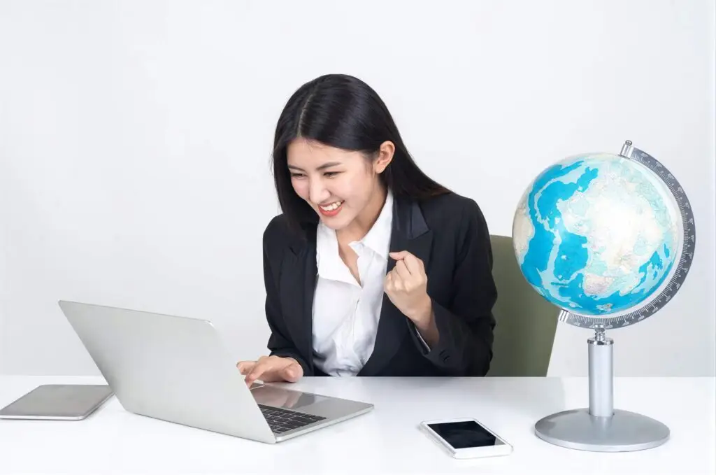 Business professional in black blazer smiling at laptop with tablet, phone, and globe on desk