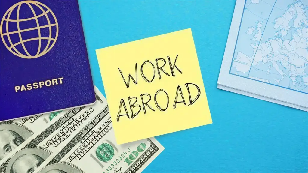 Flat lay of passport, U.S. dollars, sticky note with ‘Work Abroad,’ and map on blue background