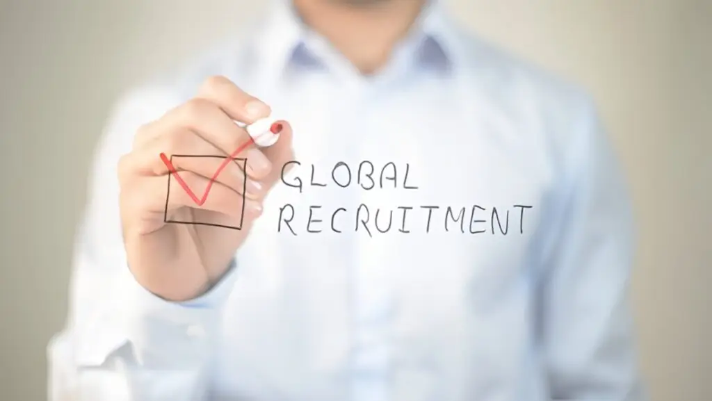 Person in white shirt writing ‘Global Recruitment’ with red checkmark on transparent surface