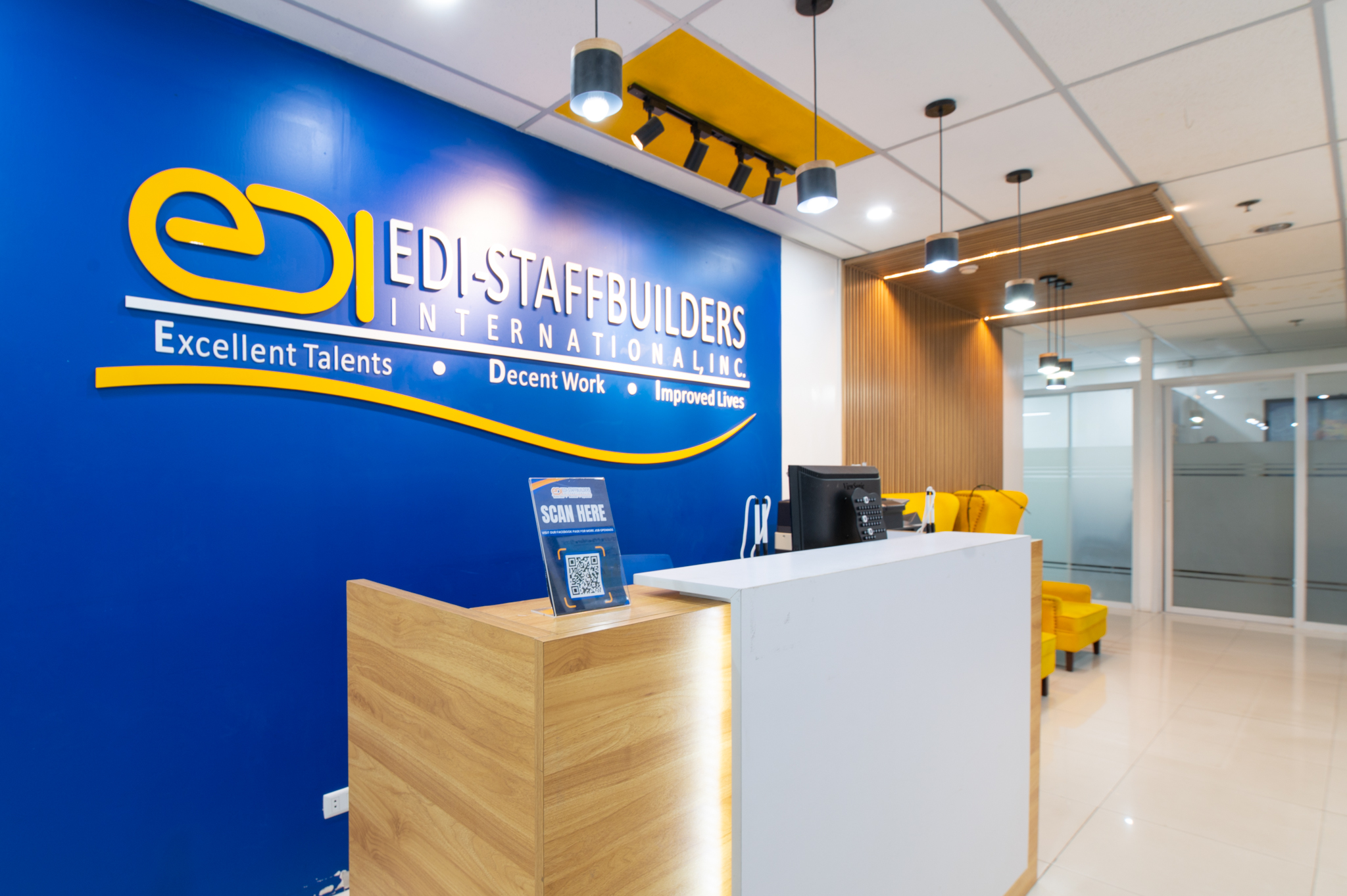 Modern office reception of EDI-STAFFBUILDERS INTERNATIONAL, INC. with a blue branded wall, white and wood reception desk, and yellow seating.