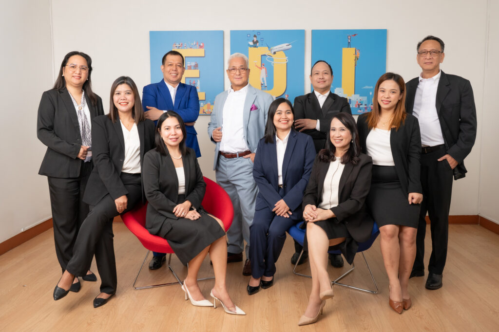 A group of 11 smiling professionals in business attire, some standing and some sitting, in front of a blue wall with large illustrated letters "EDI".